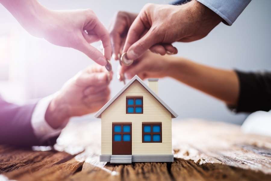 Plusieurs mains mettent en commun des pièces de monnaie dans une tirelire en forme de maison miniature sur une table en bois, symbolisant le financement d'un premier achat immobilier.